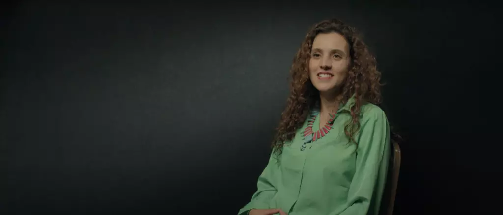 Smiling person with curly brown hair wearing a green shirt on a dark background.