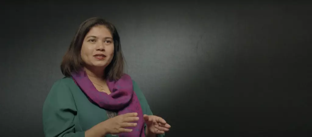 A young woman with long brown hair wearing a green top and purple scarf, standing against a dark background.