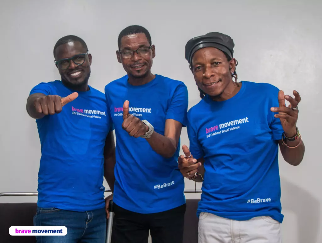 Three people wearing blue t-shirts with "Brave movement" logo, smiling and making hand gestures.